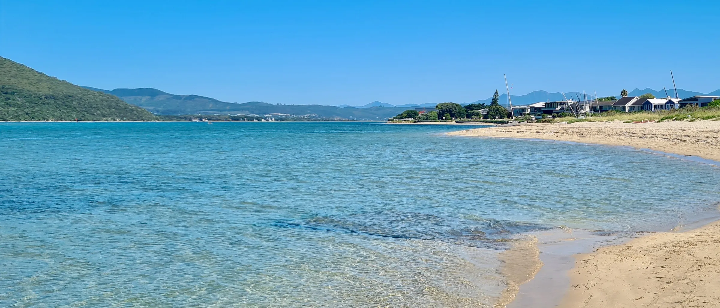 Bollard Beach on Leisure Isle: clear shallow lagoon water over pale sand, with the island's low houses and dinghy masts along the shoreline and blue mountains on the horizon.